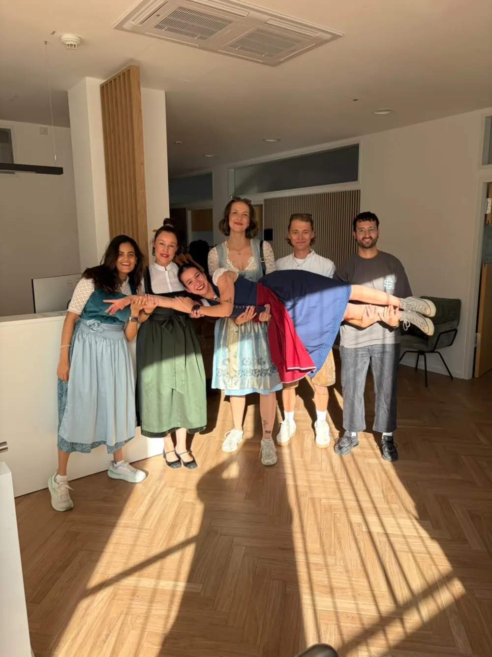 Sechs Physiotherapeutinnen und Physiotherapeuten von Eden Physiotherapie München tragen eine lachende Kollegin auf den Händen