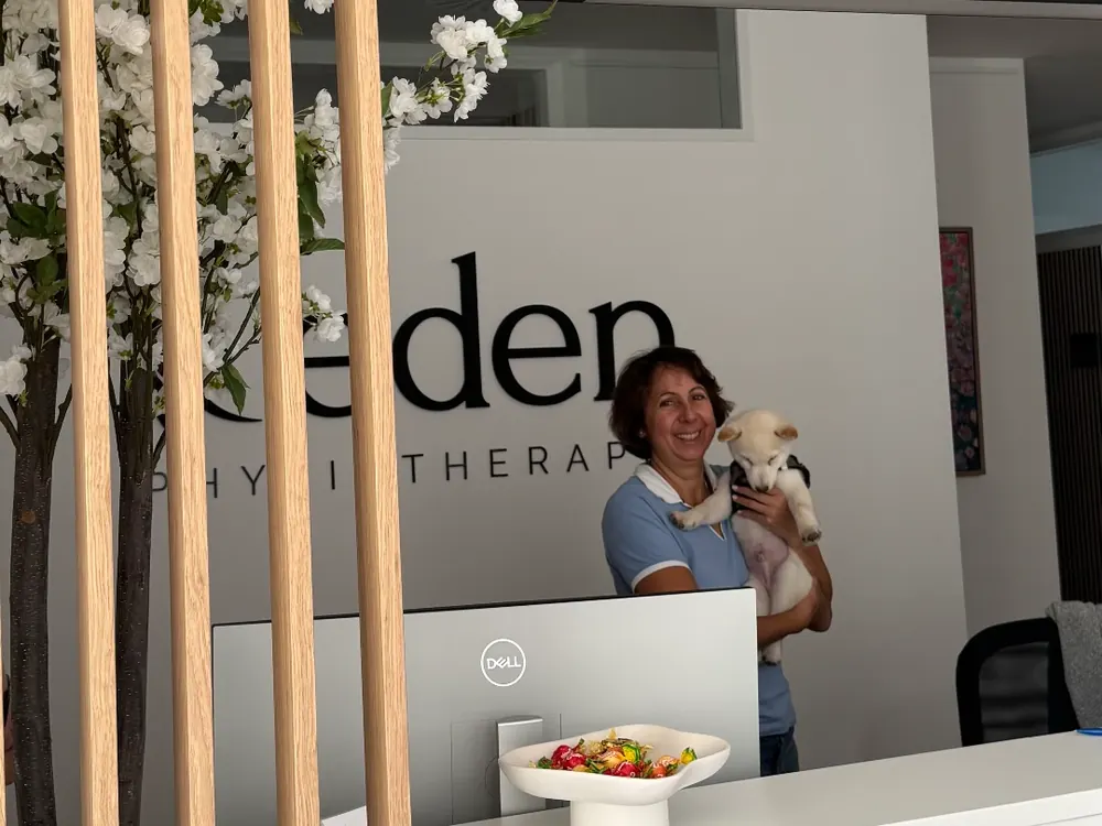 Empfang in der Praxis Eden Physiotherapie München: Eine Mitarbeiterin in hellblauem Polohemd hält einen kleinen Welpen auf dem Arm hinter dem weißen Tresen