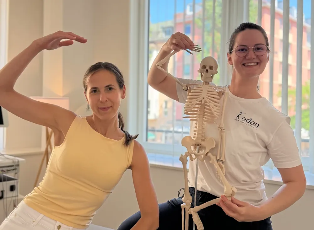 Zwei Physiotherapeutinnen von Eden Physiotherapie München posieren im Behandlungsraum mit einem anatomischen Skelettmodell – eine sitzt auf der Liege, die andere führt die Arme des Skeletts